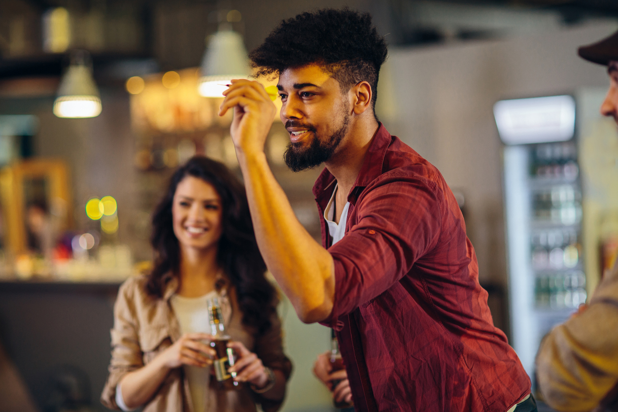 Man holding a dart at eye level, preparing to throw while friends watch and smile at a bar